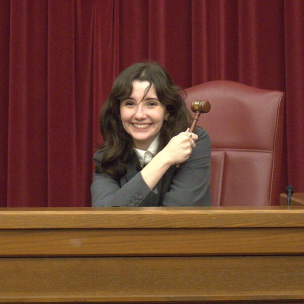 A smiling student sits at a judges bench with a gavel in their hand.