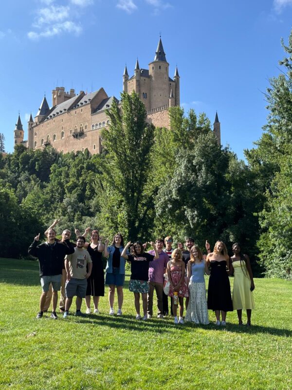 A group of people standing in front of evergreen trees with a castle-like building in the background.