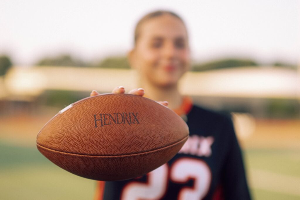 A flag football player in the background holds a football up to the camera.