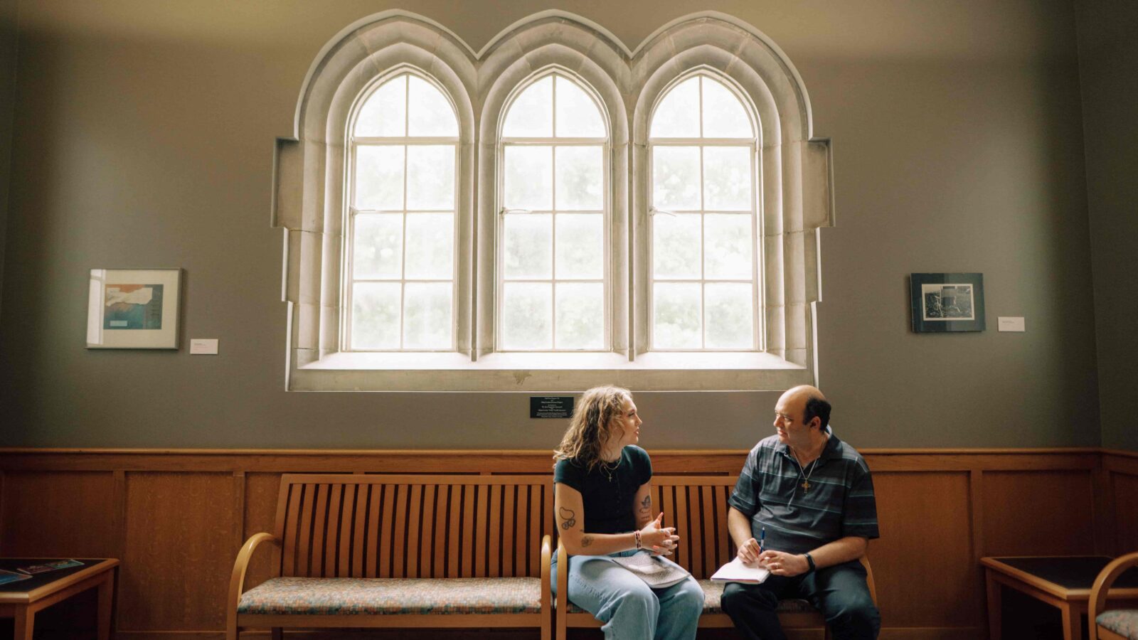 A student and mentor sit on a bench under arched windows.
