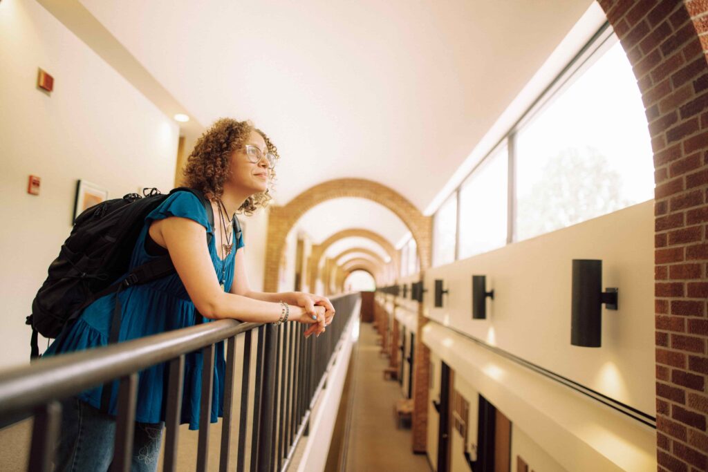 A student with a backpack leans on the banister outside a classroom in the Mills building while looking out the window.