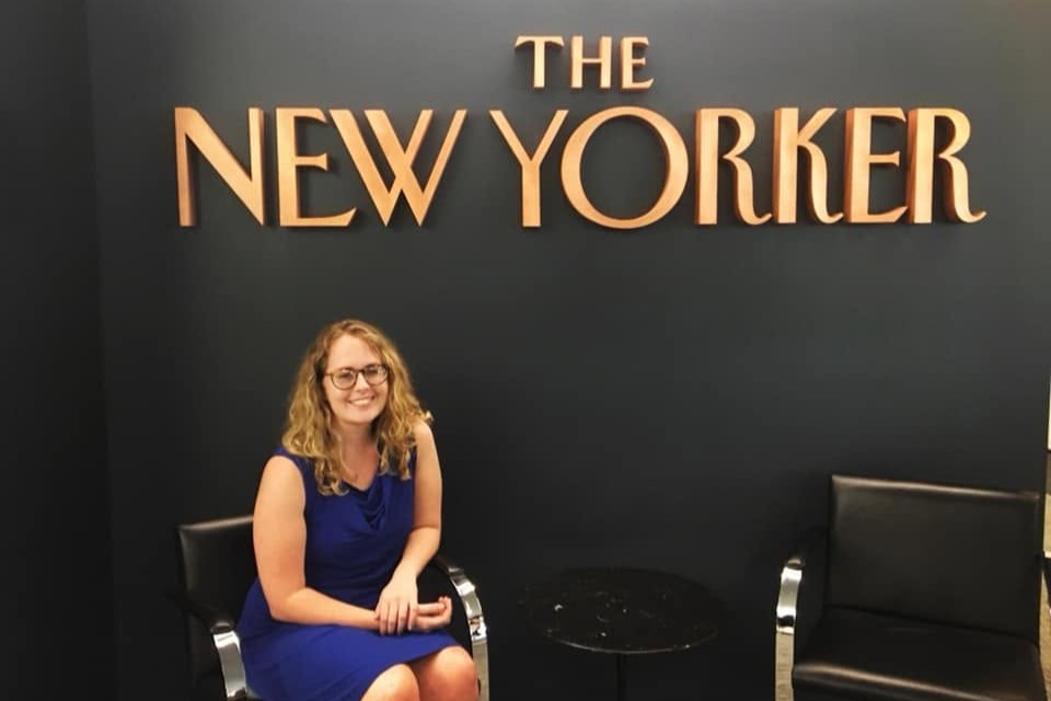 A female student wearing a blue dress smiles as she sits in a chair in front of the logo for The New Yorker.